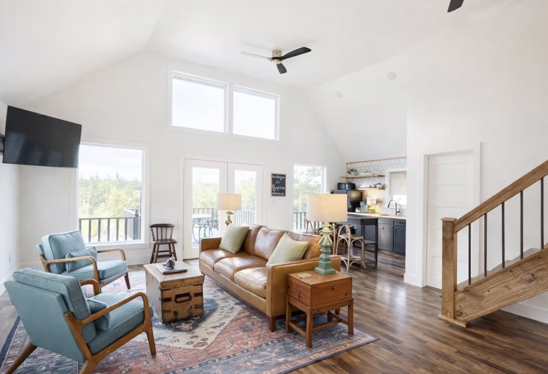 The sunlit Rockhill cabin living room with leather sofa, blue chairs, and a wooden staircase